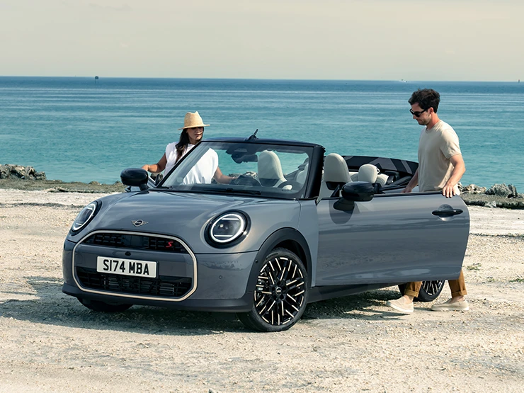 Vue avant gauche de la MINI Cooper Cabrio Copper Grey au bord d’une plage avec deux personnes à côté.