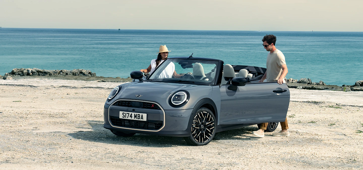 Vue avant gauche de la MINI Cooper Cabrio Copper Grey au bord d’une plage avec deux personnes à côté.