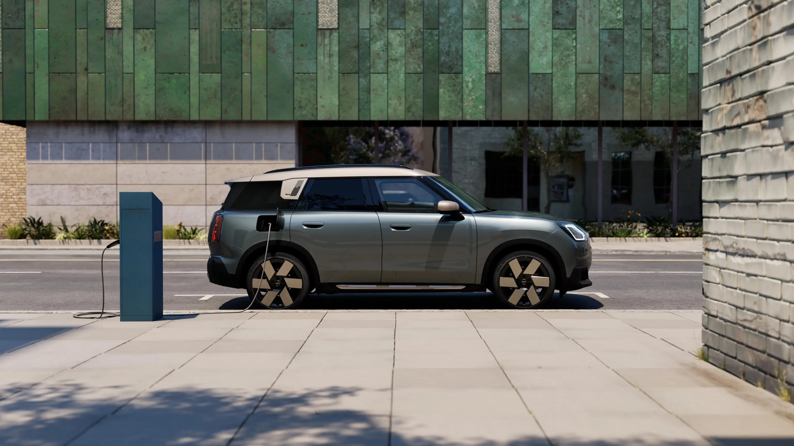 Vue latérale du MINI Countryman Electric en charge dans la rue, en Smokey Green avec toit Vibrant Silver.