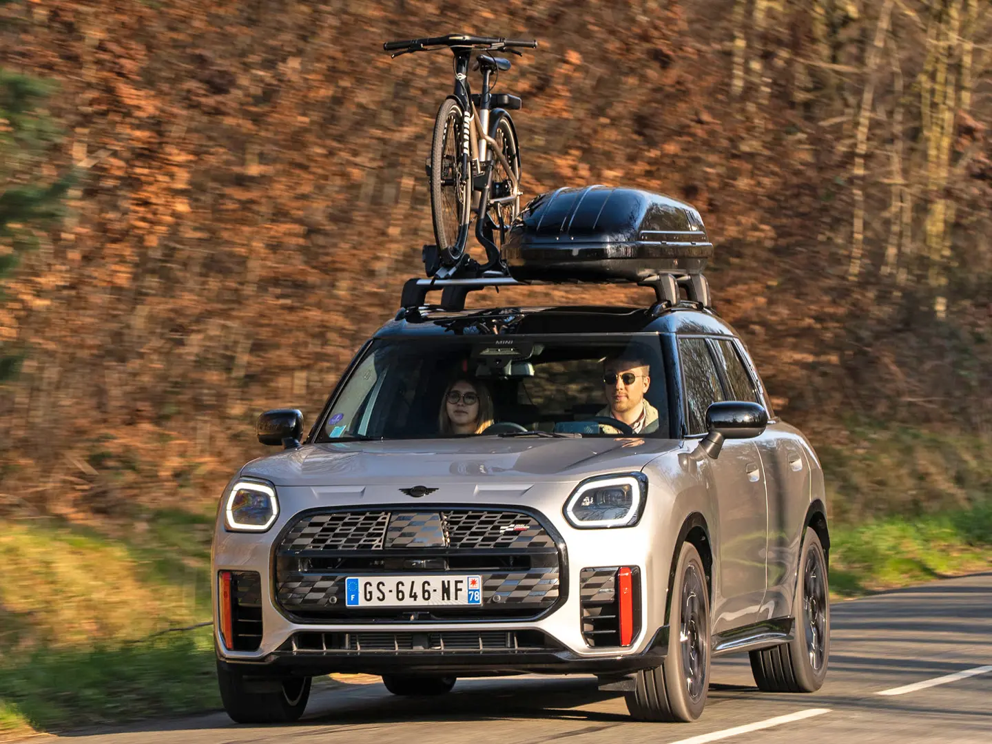 MINI Countryman Cooper en argent sur une route de montagne avec galerie de toit MINI, coffre de toit MINI, porte-vélos de toit MINI avec un vélo monté et ailette MINI John Cooper Works, vue de face. 