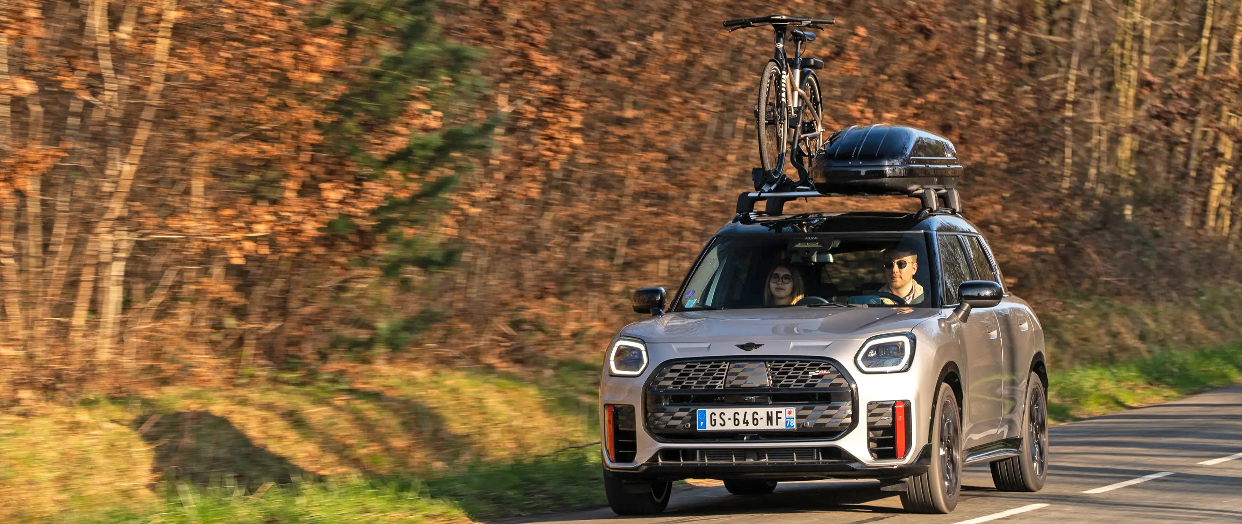 MINI Countryman Cooper en argent sur une route de montagne avec galerie de toit MINI, coffre de toit MINI, porte-vélos de toit MINI avec un vélo monté et ailette MINI John Cooper Works, vue de face. 