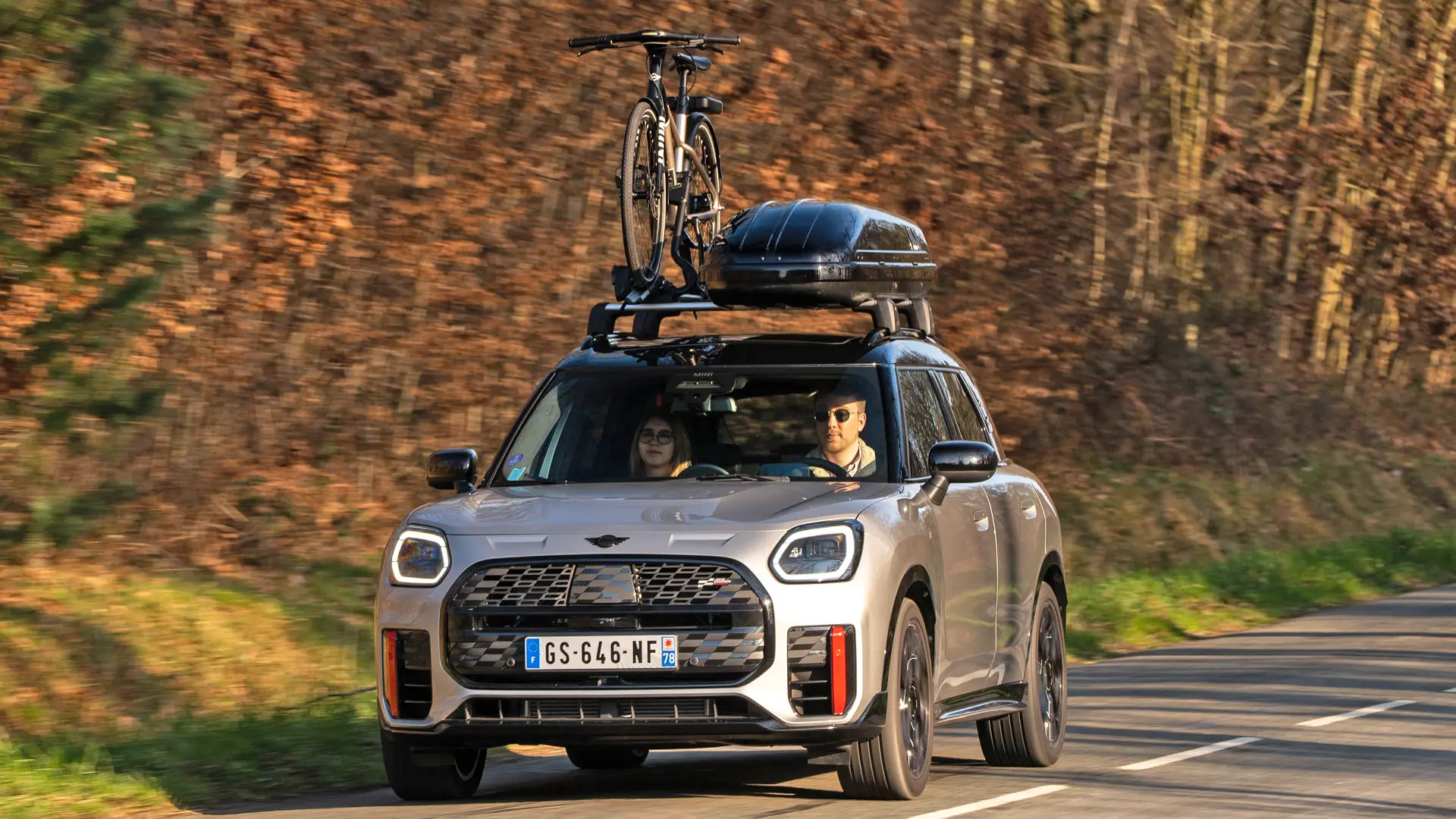 MINI Countryman Cooper en argent sur une route de montagne avec galerie de toit MINI, coffre de toit MINI, porte-vélos de toit MINI avec un vélo monté et ailette MINI John Cooper Works, vue de face. 