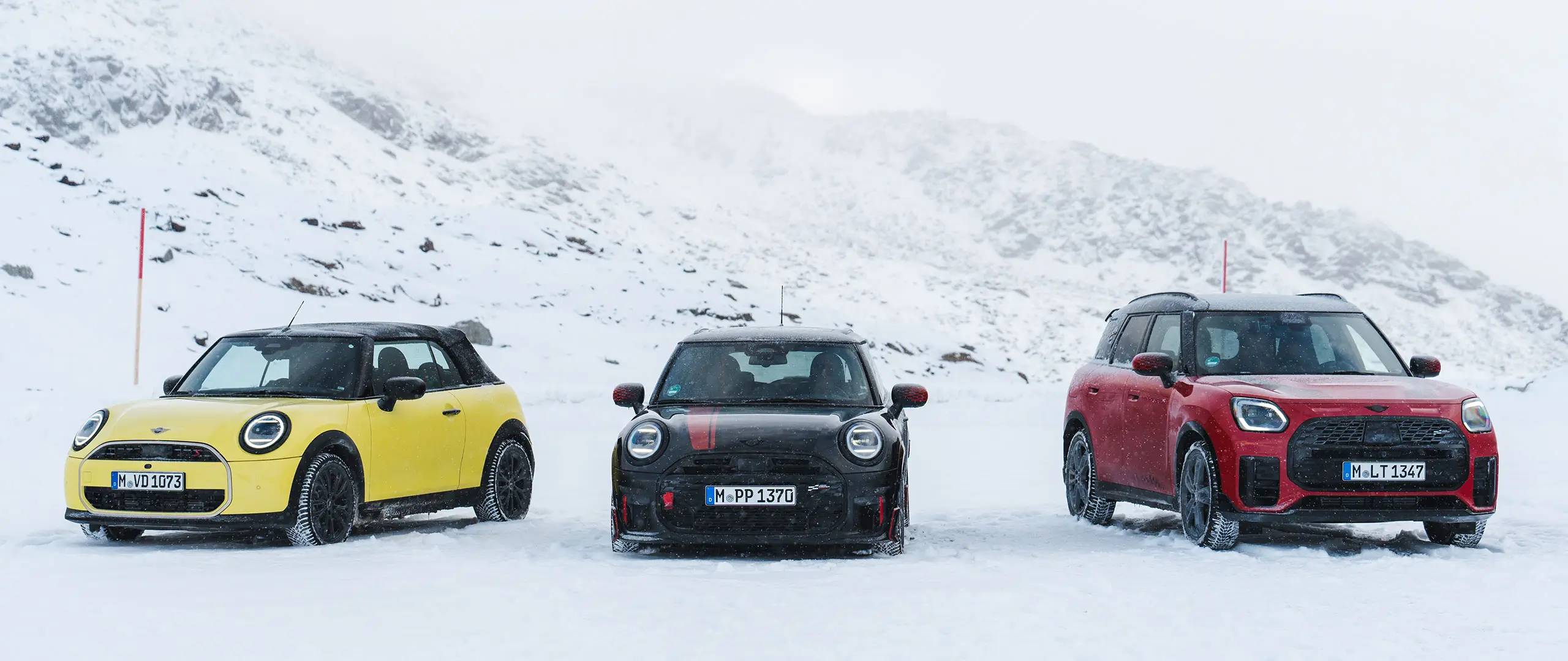 MINI Cooper Cabriolet en jaune Sunny Side, MINI John Cooper Works en Midnight Black II et MINI Countryman Cooper en Chili Red, à l’arrêt sur une route de montagne enneigée, vue de face.