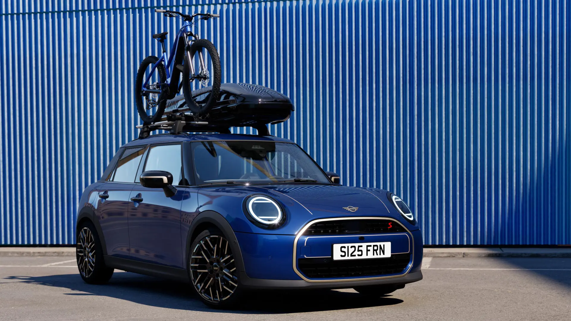 Vue avant du MINI Aceman en Indigo Sunset Blue avec porte-vélos de toit MINI, transportant un vélo de montagne et un coffre de toit MINI 320.