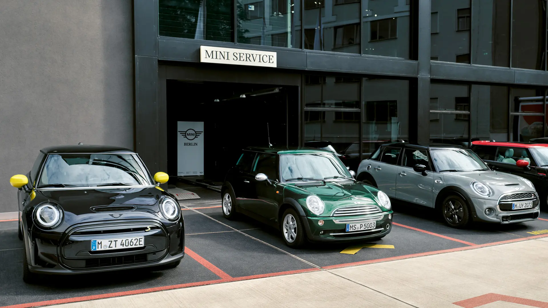 MINI Cooper Electric S noire garée devant une concession MINI, à côté d’une MINI Cooper 3 portes verte, d’une MINI 5 portes argent et d’une MINI JCW noire, vue de face. 