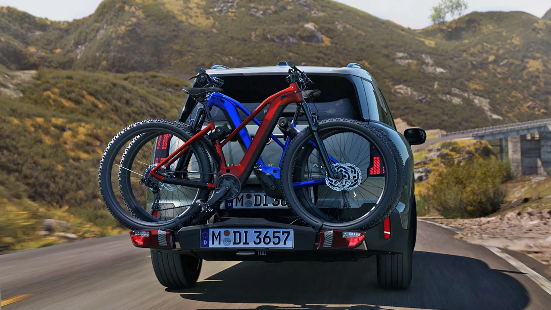 MINI Countryman Smokey Green roulant sur une route de montagne, dotée d’un porte-vélos arrière transportant deux VTT, vue arrière.